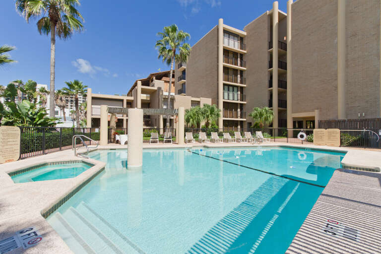 Large resort-style swimming pool and hot tub surrounded by palm trees and lounge chairs, perfect for relaxing on South Padre Island.