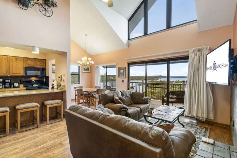 Vacation Rental Living Area With Leather Sofa, Mounted TV, And Kitchen Bar, Looking Out To Lake View Through Large Windows Vacation Rental Living Area With Leather Sofa, Mounted TV, And Kitchen Bar, Looking Out To Lake View Through Large Windows