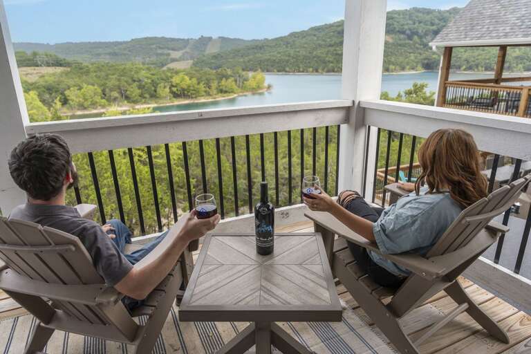 Two People Relaxing, Sipping Drinks, Overlooking A Lush Lake Landscape From A Balcony