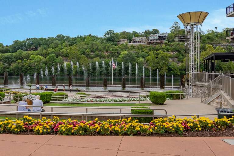 Riverside Plaza With Rows Of Fountains, Vibrant Flowerbeds, And Hillside Homes In Background