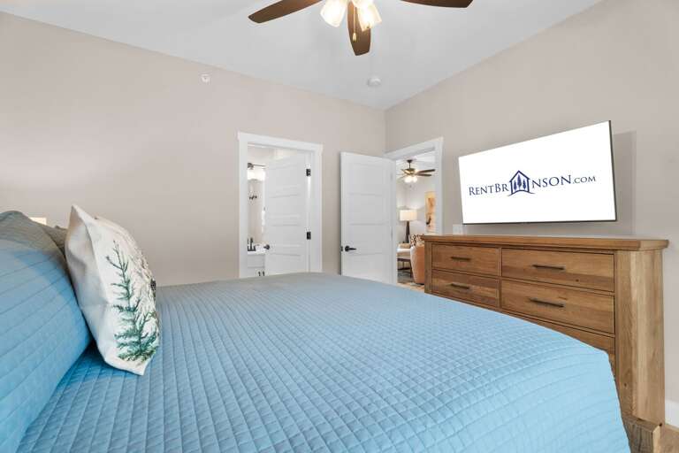 Bedroom With Blue Bedding, Wooden Dresser, Ceiling Fan, TV Displaying Logo, And Open Doors