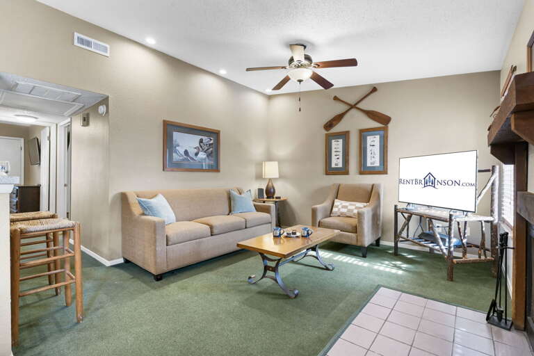 Living Space With Beige Walls, Green Carpet, And Furnishings Including Sofas, Coffee Table, And A Ceiling Fan