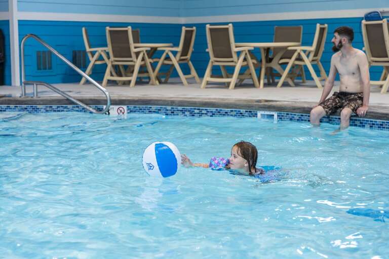 Child Chasing Ball In Pool, Man Watching, Poolside Loungers Visible