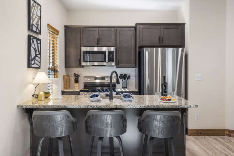 Compact Kitchen With Stools, Stainless Steel Appliances, And Sleek Stone Countertop