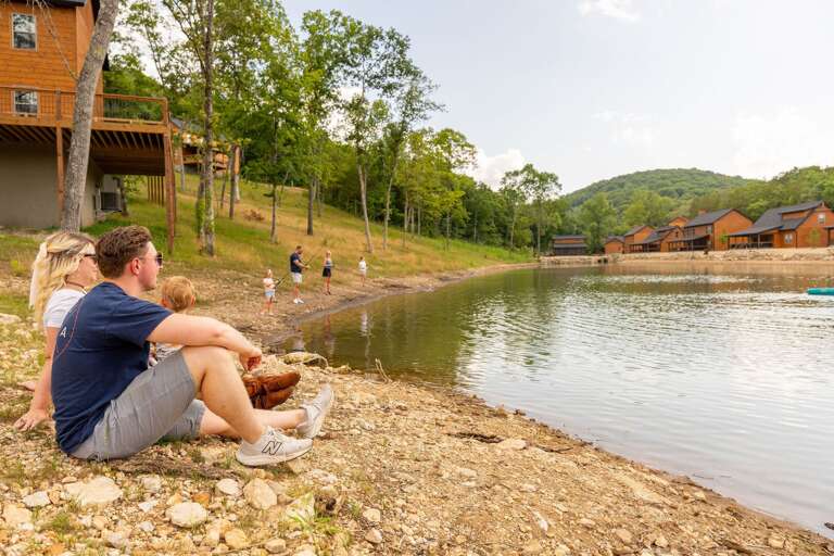 Family By The Lakeside, Lounging And Looking At Water With Wooden Structures On Hill