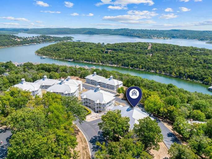 Aerial View Of A Sprawling Riverfront Property With Multiple Distinctively Designed Structures, Surrounded By Lush Trees And Overlooking A Winding River