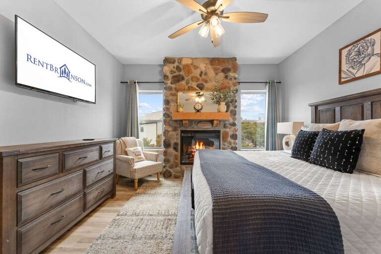 Bedroom With Stone Fireplace, Ceiling Fan, Wooden Dresser, And Plush Bed