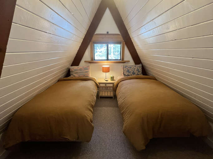 Drift off in this cozy A-frame loft, where twin beds frame a peaceful forest view through the window. Soft lighting and rustic charm create your perfect retreat.