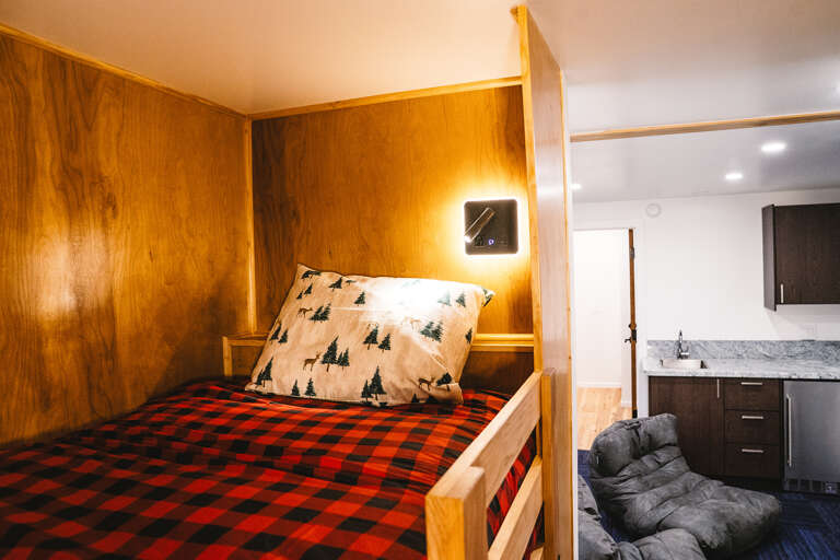 Cozy up in your rustic wood-paneled bunk with soft red plaid bedding and charming forest-print pillows for mountain cabin dreams.