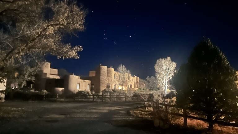 Adobe-style buildings create a serene desert retreat under starlit winter skies, surrounded by snow-dusted trees and natural stone landscaping.