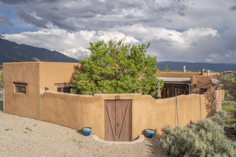 Adobe-style entrance with handcrafted wooden gate and lush greenery, set beneath dramatic mountain skies.