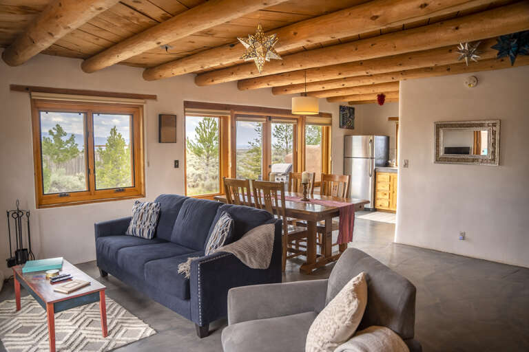 Open living space with authentic log-beam ceilings and large windows framing views of natural scenery. Cozy seating invites you to unwind while the adjoining dining area sets the stage for memorable meals together.