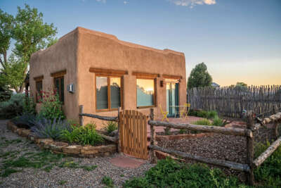 Charming adobe-style casita nestled in a thoughtfully landscaped garden, framed by rustic fencing and native plants. Warm earthy tones and traditional Southwestern architecture create a welcoming retreat.