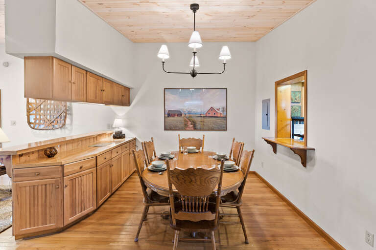 Wooden Dining Table Flanked By Kitchen, Chandelier Overhead