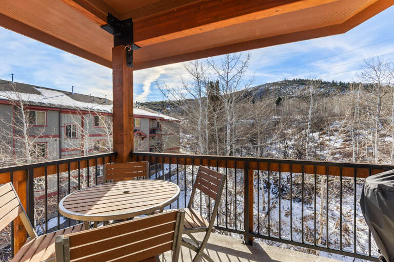 A cozy covered balcony offering mountain and neighborhood views, with a small table and chairs for relaxing mornings and wintry alpine scenery just beyond your door.