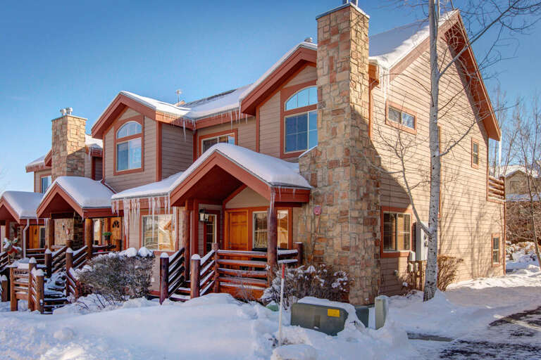 A charming mountain-style property featuring stone chimneys and warm wood siding, nestled in a snowy neighborhood with clear winter skies overhead.