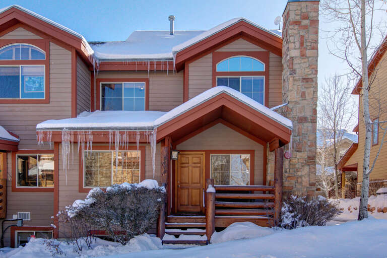 A charming mountain-style townhome nestled in a snowy neighborhood, featuring warm wood accents, a stone chimney, and welcoming front entrance framed by winter scenery.