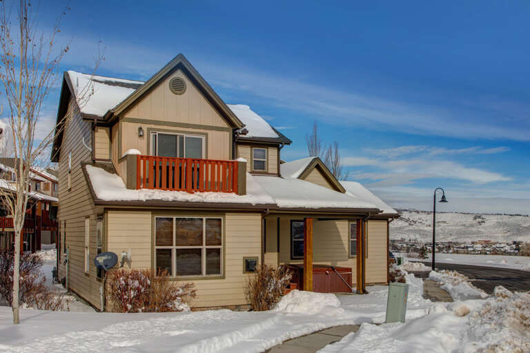 A charming two-story home with warm tan siding and cheerful orange accents nestled in a snowy mountain neighborhood, offering peaceful winter surroundings.