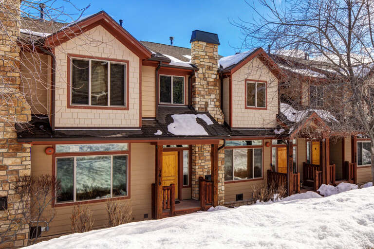 A charming alpine-style property with stone and wood details, nestled in a snowy mountain setting under clear blue skies, offering a warm welcome for your winter retreat.