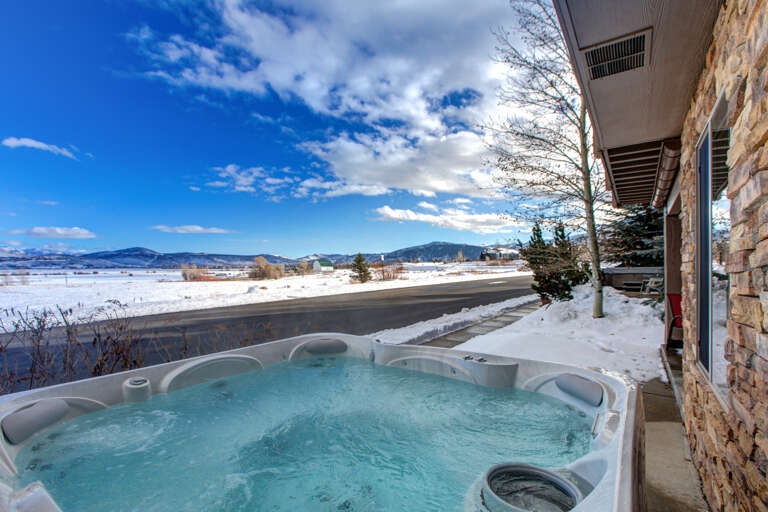 A hot tub overlooking a snowy landscape and distant mountains, situated on a covered terrace adjacent to a stone-accented building along a quiet street.