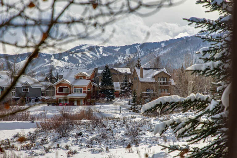 A quiet winter neighborhood framed by snow-dusted pines and evergreens, nestled below mountain slopes dotted with ski runs.