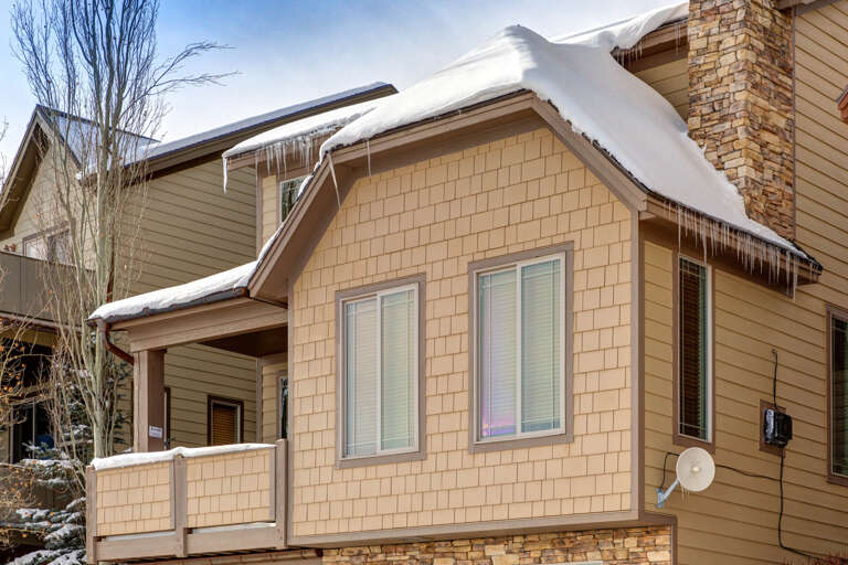A cozy mountain retreat awaits, its shingled exterior and stone accents blending beautifully with the snowy landscape. Icicles glisten along the roofline under crisp winter skies.