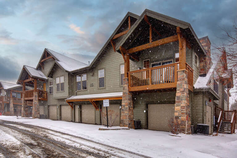 Mountain townhome complex with covered porches and garage parking, set against a snowy winter backdrop with dramatic evening skies.