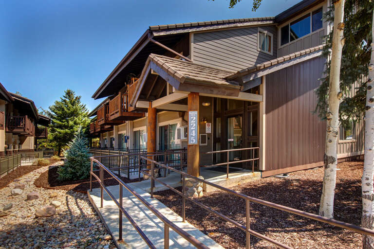 Modern mountain retreat with accessible ramps, surrounded by native evergreens and aspens under a clear blue sky. A tranquil arrival setting with thoughtful landscaping and convenient ground-level access.