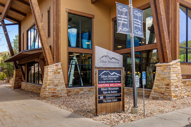 Silver Mountain Sports Club & Spa entrance features rustic mountain architecture with stone pillars, timber beams, and large windows. The facility includes spa services, fitness amenities, and a kids cabin.