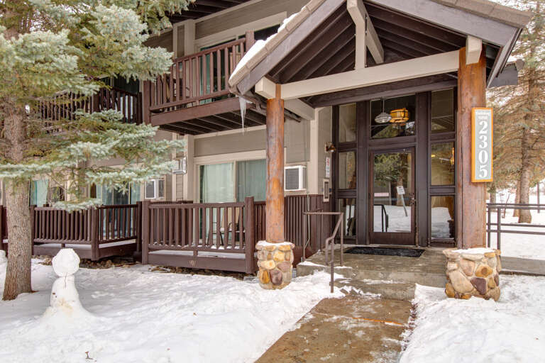 A charming mountain retreat nestled among snow-dusted pines, featuring rustic wood beams, stone accents, and a welcoming entry with balconies to enjoy the alpine setting.