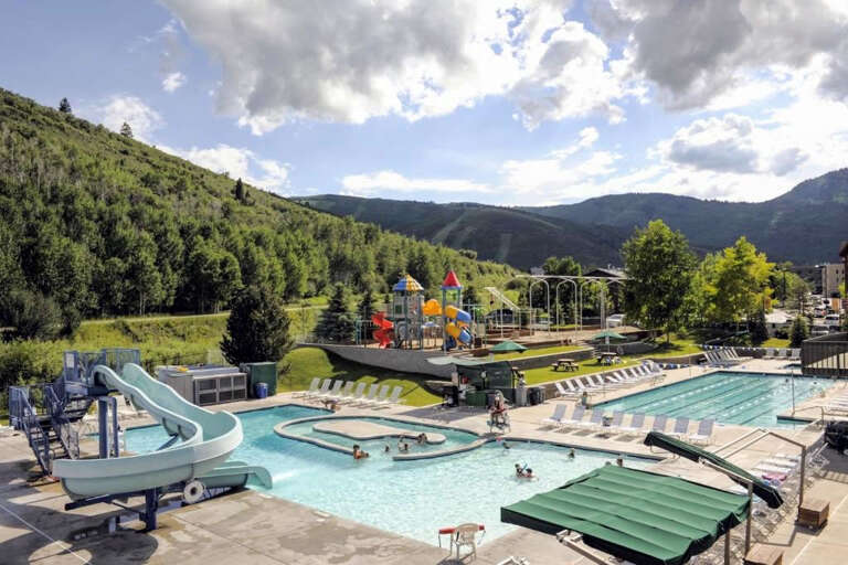 Mountain resort aquatic center set amid forested slopes, featuring water slides, lap pool, lazy river, and colorful playground under open skies.