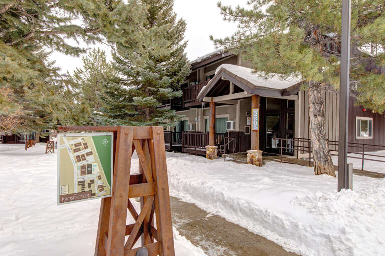 A rustic mountain lodge entrance nestled among tall pines, with snow-dusted grounds and natural wood details creating a welcoming winter retreat atmosphere.