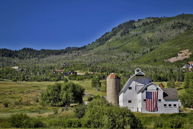 The property sits in a picturesque valley surrounded by lush forested hillsides, with a charming white barn and vintage silo marking the entrance to the area.