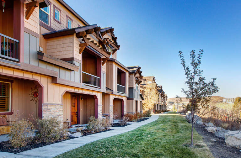 A row of modern residential units features private entrances, balconies, and welcoming walkways amid beautifully landscaped grounds with young trees and stone accents under clear skies.