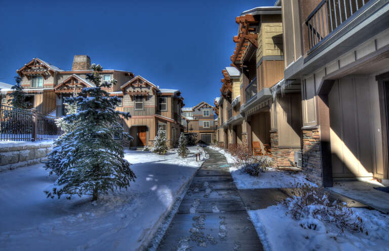 A charming winter scene showcases snow-dusted alpine-style buildings under a crisp blue sky, inviting guests to a cozy mountain retreat surrounded by evergreens and peaceful pathways.