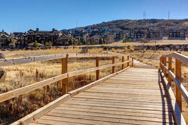 A scenic wooden boardwalk winds through natural grassland near the property, with mountain resort buildings visible in the background under clear blue skies—a tranquil area for nature walks.