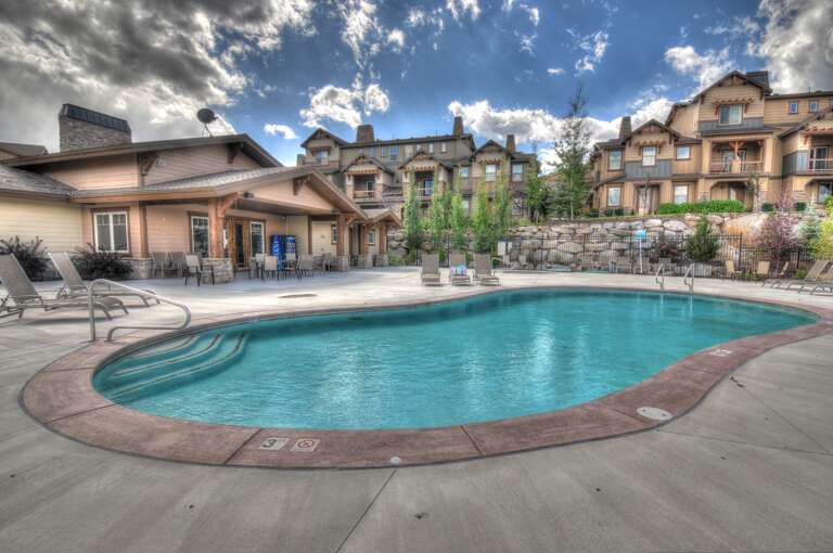 A shared outdoor pool with curved steps and sundeck surrounded by mountain lodge-style buildings, native landscaping, and open sky—part of the property's communal amenities.