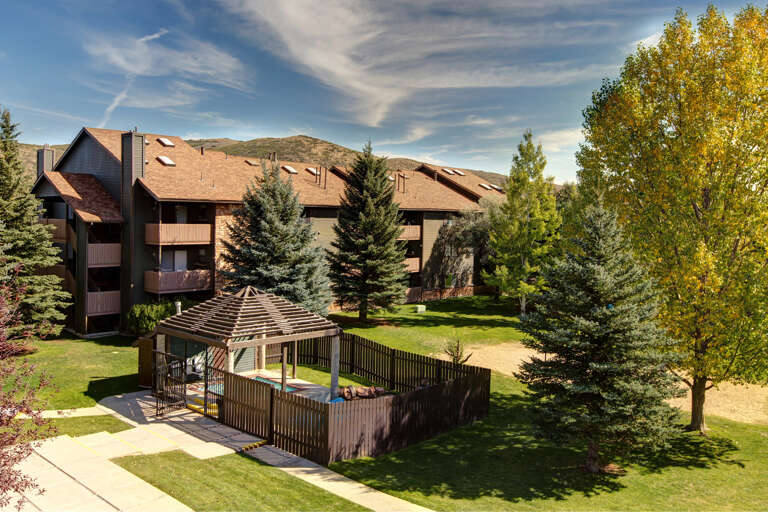 Multi-level vacation condo complex nestled in a scenic mountain valley, featuring manicured lawns, mature evergreen and aspen trees, and a covered hot tub gazebo surrounded by seasonal foliage.
