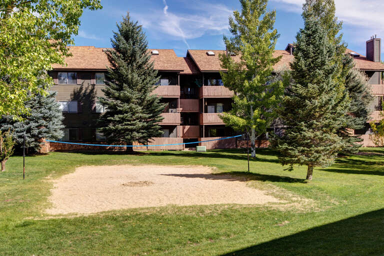 The property features a volleyball court set in a spacious, well-maintained lawn surrounded by mature evergreen and deciduous trees, with the multi-level building providing a scenic mountain retreat backdrop.