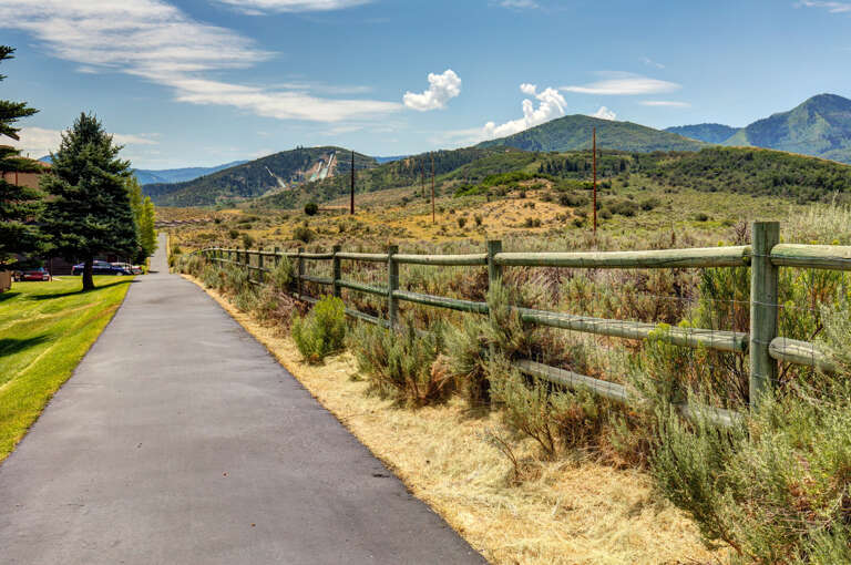A paved pathway lines the property grounds, bordered by rustic fencing and open terrain leading to panoramic mountain views under wide blue skies.