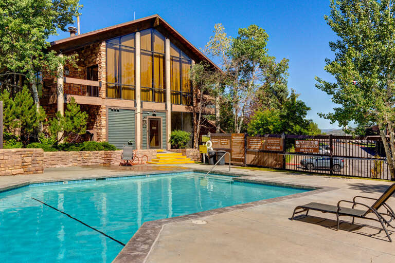 Community pool set in a landscaped courtyard with modern two-story building featuring floor-to-ceiling windows, sunlit deck, and mountain views framed by mature trees.