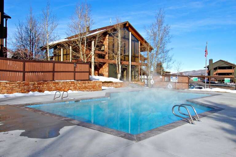 This resort features a heated outdoor pool and hot tub area set against a stunning mountain backdrop, with modern ski lodge architecture and winter scenery visible throughout the property.