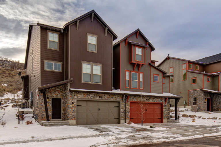 Modern multi-unit residential buildings featuring stone and siding exteriors with attached garages, set in a hillside neighborhood with winter landscaping.