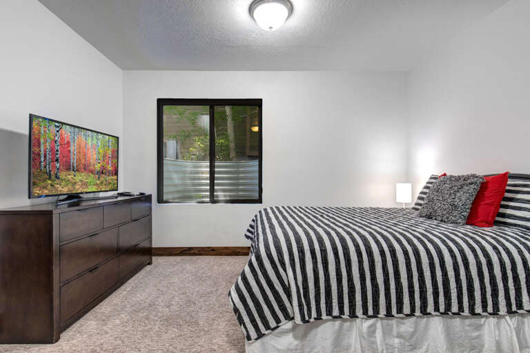 Your cozy bedroom features a plush bed with bold striped linens and pops of red, a large TV for movie nights, and a peaceful window view to wake up to each morning.