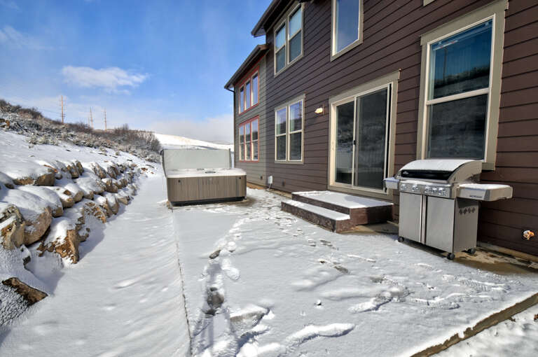 A snow-dusted patio featuring a private hot tub and BBQ grill, backed by scenic hillside views—perfect for year-round outdoor relaxation and mountain getaways.