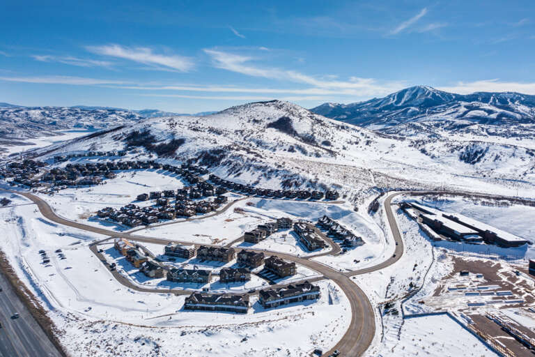 Aerial view of a mountain resort community nestled in snow-covered hills, with ski slopes visible in the distance under a brilliant blue sky.