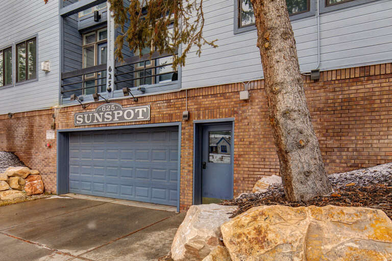 The Sunspot property entrance features a two-car garage with direct access and a welcoming side door, set against natural landscaping with mature trees and stonework.