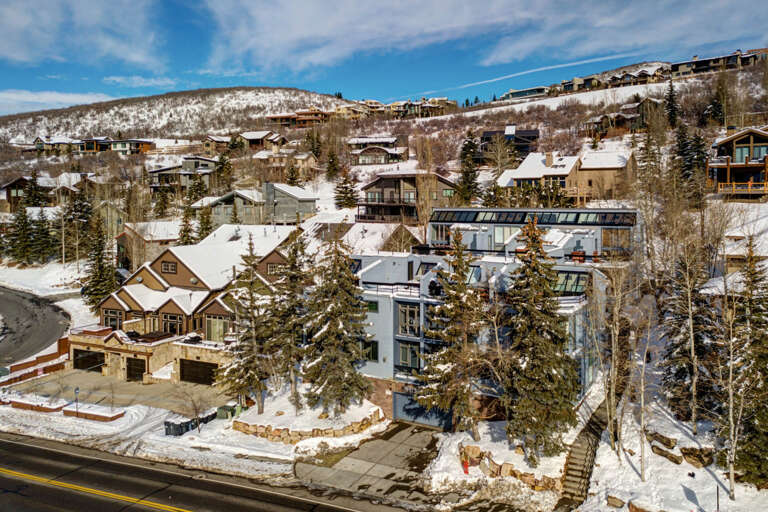 An alpine ski village nestled in snow-covered mountains, showcasing a scenic winter neighborhood with charming lodges, evergreen trees, and hillside homes beneath a crisp blue sky.