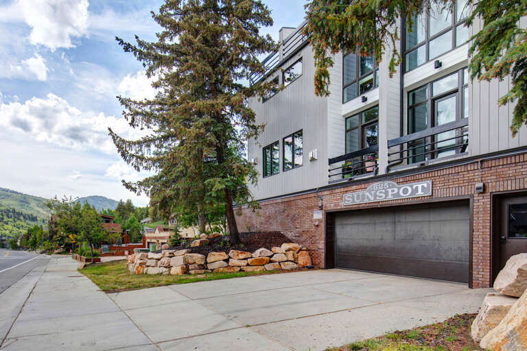 Modern mountain property entrance with natural landscaping and convenient parking, set against scenic hillside backdrop.