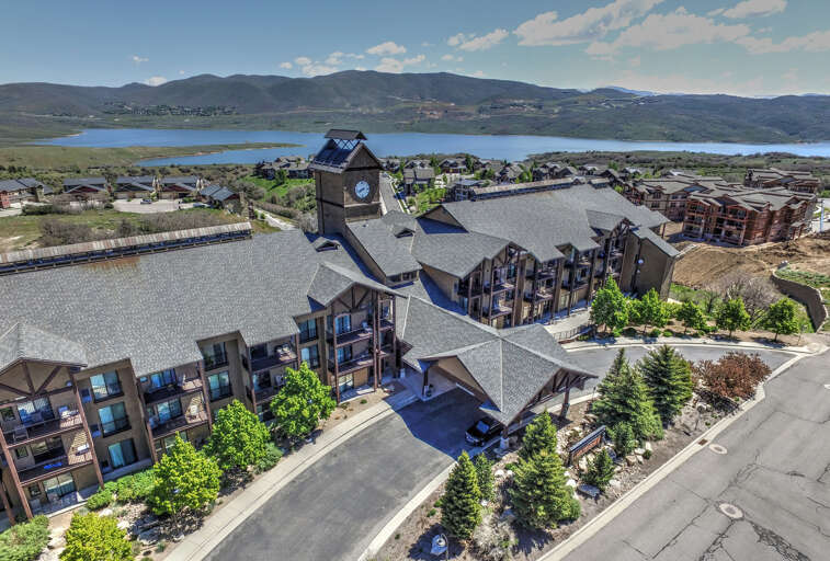 Aerial view of the property complex featuring distinctive clock tower architecture, situated near a scenic lake with mountain views in the surrounding landscape.